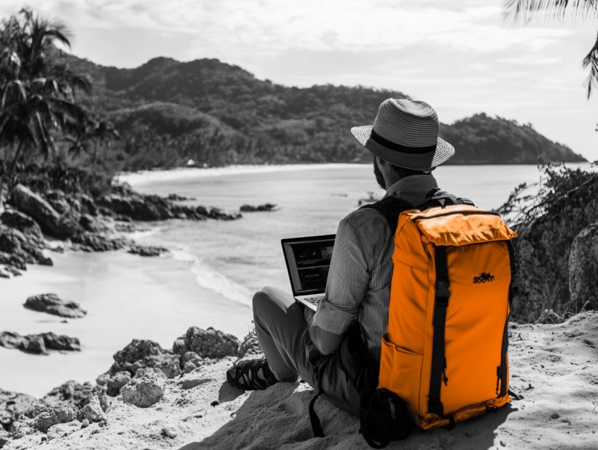 Ein Mann mit Hut sitzt auf einer Klippe und schaut zu Meer. Er hat einen Hut auf, einen orangenen Rucksack auf dem Rücken und arbeitet am Laptop. Frau zieht zwei orangene Koffer hinter sich her, LLC Gründung, Solution Thirteen LLC, Firma im Ausland gründen